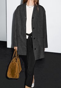 Femme portant un manteau gris foncé boutonné, un haut blanc, un pantalon noir, portant un grand sac à main en daim marron, marchant sur un sol sombre à l’intérieur.