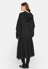 Czarna, oversize bluza z kapturem, zestawiona z warstwową czarną spódnicą z tiulu. Czarne dzianinowe ocieplacze na nogi i płaskie buty dopełniają całość.