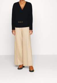 Suéter negro de punto con escote en V y detalle dorado en la cintura, combinado con pantalones de pierna ancha beige y mocasines marrones.