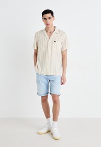 Cămașă cu mânecă scurtă, de culoare bej deschis, cu dungi verticale, asortată cu șorturi din denim bleu deschis, sosete albe și adidași casual albi.