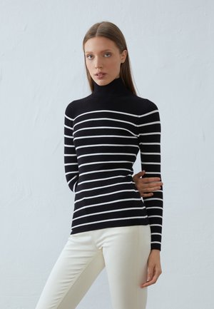 Pull à col roulé noir et blanc rayé avec une texture côtelée. Caractérisé par des manches longues et un design ajusté, associé à un pantalon de couleur crème.
