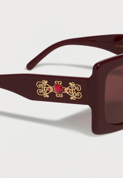 Lunettes de soleil bordeaux foncé avec montures carrées comportant un détail ornemental doré et une pierre précieuse rouge sur la branche.