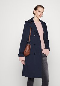 Trench coat de marin, cu design cu două rânduri de nasturi, nasturi mari, epauleți pe umăr și talie cu curea, purtat peste un pulover roz deschis și blugi gri.