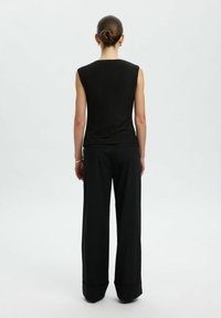 Donna con i capelli raccolti in un bun, che indossa un top senza maniche nero e pantaloni larghi neri, in piedi di spalle contro uno sfondo neutro.