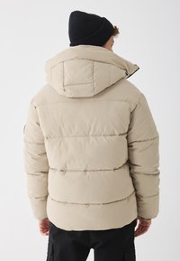 Jachetă puffer bej cu glugă, având panouri orizontale cusute și o textură netedă, asociată cu o căciulă din tricot neagră.