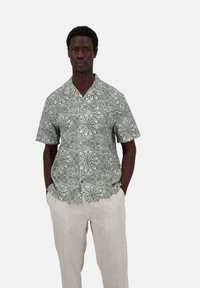 Camicia a maniche corte verde con un motivo floreale bianco; tessuto leggero; design con colletto; bottoni sul davanti; abbinata a pantaloni di lino chiari.
