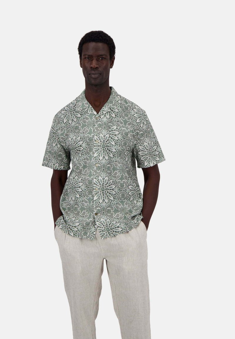 Camicia a maniche corte verde con un motivo floreale bianco; tessuto leggero; design con colletto; bottoni sul davanti; abbinata a pantaloni di lino chiari.