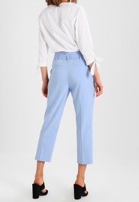 Vaaleansiniset mittatilaustyönä tehdyt housut, joissa on korkea vyötärö, lyhyt pituus ja sivutaskut, yhdistettynä valkoiseen paitaankin, jossa on sidottavat hihat.