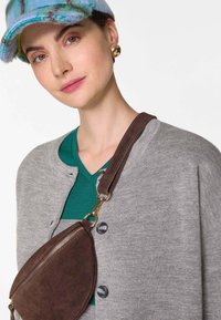 Grau gestrickte Jacke mit Knopfschluss, grünes V-Ausschnitt-Shirt und eine braune Wildleder-Crossbody-Tasche mit Reißverschluss, kombiniert mit einer mehrfarbigen Mütze.