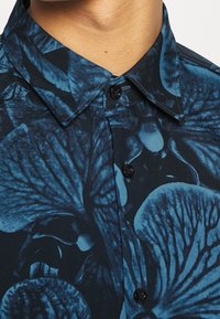 Korte mouw shirt in marineblauw met blauw bloemenpatroon. Heeft een button-down kraag en zwarte knopen. Gladde stoftextuur.