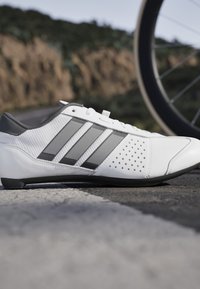 Bílé a šedé cyklistické boty na dlažbě s částečně viditelným kolem a rozmazanou přírodní krajinou v pozadí.