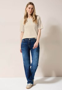 Beige tričko s krátkým rukávem v kombinaci s modrými džínami. Džíny mají zapínání na knoflík a mírné vyblednutí. Modelka stojí na světlém pozadí.