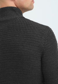 Mørkegrå strikket sweater med høj krave, der har et struktureret mønster og en tætsiddende design, der viser ribbet detaljering ved halsudskæringen.