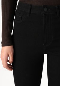 Μαύρα τζιν παντελόνια με ψηλή μέση, που διαθέτουν μπροστινή τσέπη, κλείσιμο με κουμπί και διακριτές ραφές. Στενή εφαρμογή με λείες υφές.