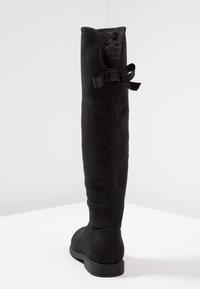 Czarny zamszowy kozak do kolan z płaską podeszwą i dekoracyjną kokardą z tyłu, wyeksponowany na tle białego tła.