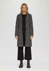 Frau steht da und trägt einen grauen karierten langen Mantel über einem schwarzen Pullover und weit geschnittenen schwarzen Hosen mit schwarzen Stiefeletten vor einem weißen Hintergrund.