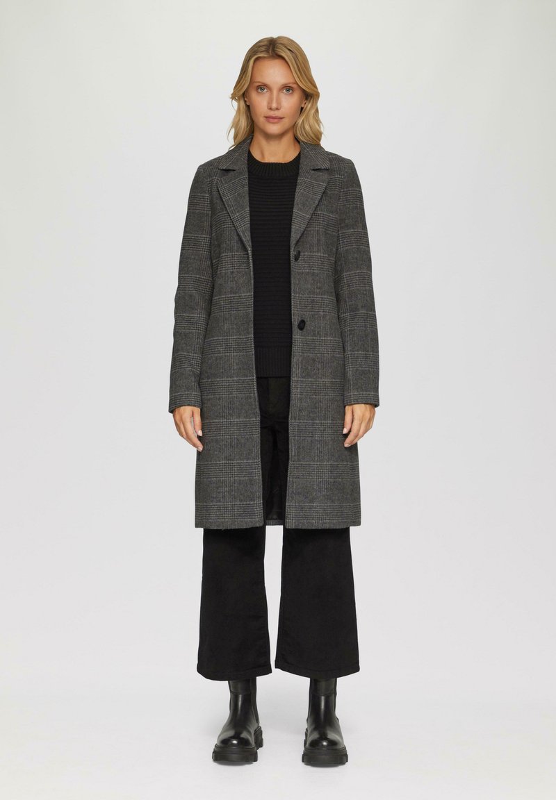 Femme se tenant debout portant un long manteau gris à carreaux sur un pull noir et un pantalon large noir avec des bottines noires sur fond blanc.