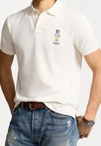 Vit polo-shirt med en björngrafik och texten "POLO". Kombinerad med slitna blå jeans och ett brunt läderbälte, vilket visar en klassisk passform.