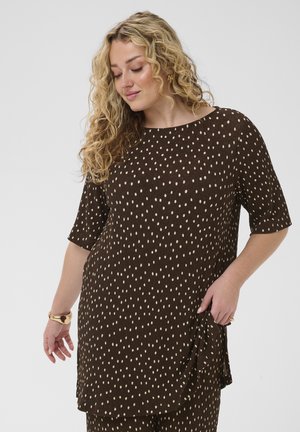 Femme aux cheveux blonds bouclés, portant une robe marron à pois blancs, tenant le tissu de la robe et portant un bracelet en or et des boucles d'oreilles.