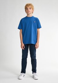 Camiseta de algodón azul con cuello redondo y mangas cortas. Presenta un pequeño parche con logotipo en el lado izquierdo del pecho. Combinada con jeans oscuros y zapatillas blancas.