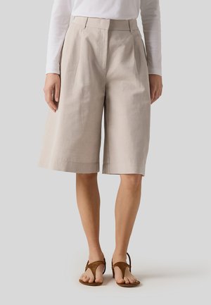 Kvinde iført beige vidbenede shorts til knæet, hvid langærmet top og brune flade sandaler, stående mod en ensfarvet baggrund.