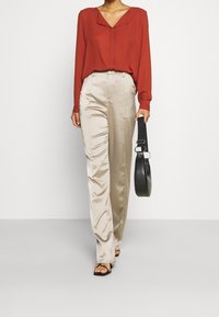 Blouse rouge à manches longues avec un décolleté en V, associée à un pantalon beige en satin taille haute. Un sac à main noir et des sandales plates viennent parfaire le look.