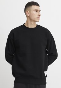 Sudadera negra con cuello redondo, hombros caídos, puños acanalados y dobladillo. Presenta un detalle de etiqueta blanca en el lado inferior.