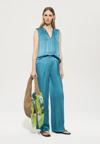 Türkisfarbener Satin-Top ohne Ärmel und weit geschnittene Hose, kombiniert mit einer geflochtenen Totetasche und einem grün gemusterten Schal. Flache Sandalen runden den Look ab.