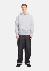 Grauer Baumwoll-Hoodie mit Kordelzugkapuze, Fronttasche und Logodetail, kombiniert mit locker sitzenden dunklen Jeans und schwarzen Schuhen.