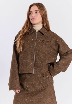 Femme portant une veste zippée marron mouchetée et une jupe assortie avec un col roulé beige, posant avec une main sur la hanche.