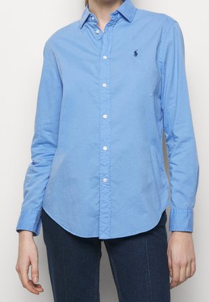 Osoba ubrana w dopasowaną jasnoniebieską koszulę na guziki z długimi rękawami oraz ciemnoniebieskie dżinsy, stojąca na jednolitym tle.