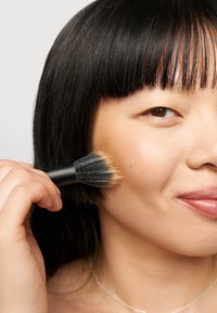 Sminkebørste med svart håndtak og myke bust, som påføres kinnet og viser en jevn hudtekstur og naturlig finish.