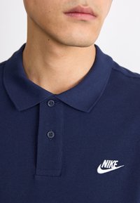Námornícka modrá polo košeľa z texturovanej látky, s golierom, dvoma gombíkmi a bielym logom Nike na ľavej strane.