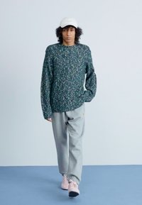 Trui in donkergroen met een gestructureerd bloemenpatroon, grijze joggingbroek en lichtroze schoenen. Het model draagt een witte pet en staat tegen een effen achtergrond.