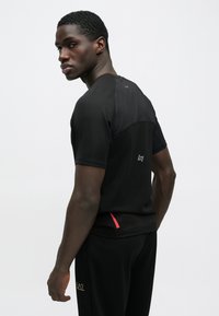 Melna sporta T-krekls ar īsām piedurknēm, teksturētu audumu, kontrastējošām smalkām detaļām uz pleciem un apakšmalu, kā arī minimālu zīmola elementu aizmugurē.