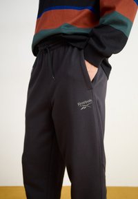 Crne trenerke od mekog materijala, s elastičnim pojasom i vezicom, bočnim džepovima i Reebok logom na butu.