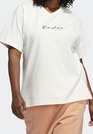 Person iført en off-white V-hals kortærmet T-shirt med teksten "Rodeo" og lyse ferskenfarvede bukser, stående mod en ensfarvet baggrund.