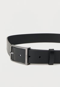 Μαύρη δερμάτινη ζώνη με λεία υφή, που διαθέτει ασημένια ορθογώνια αγκράφα και πολλές τρύπες ρύθμισης κατά μήκος του ιμάντα.