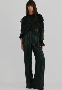 Černá průhledná blůza s volánky a vysokým límcem, spárovaná s tmavě zelenými kalhotami s vertikálním pruhováním a vysokým pasem. Oba kousky mají splývavý design.