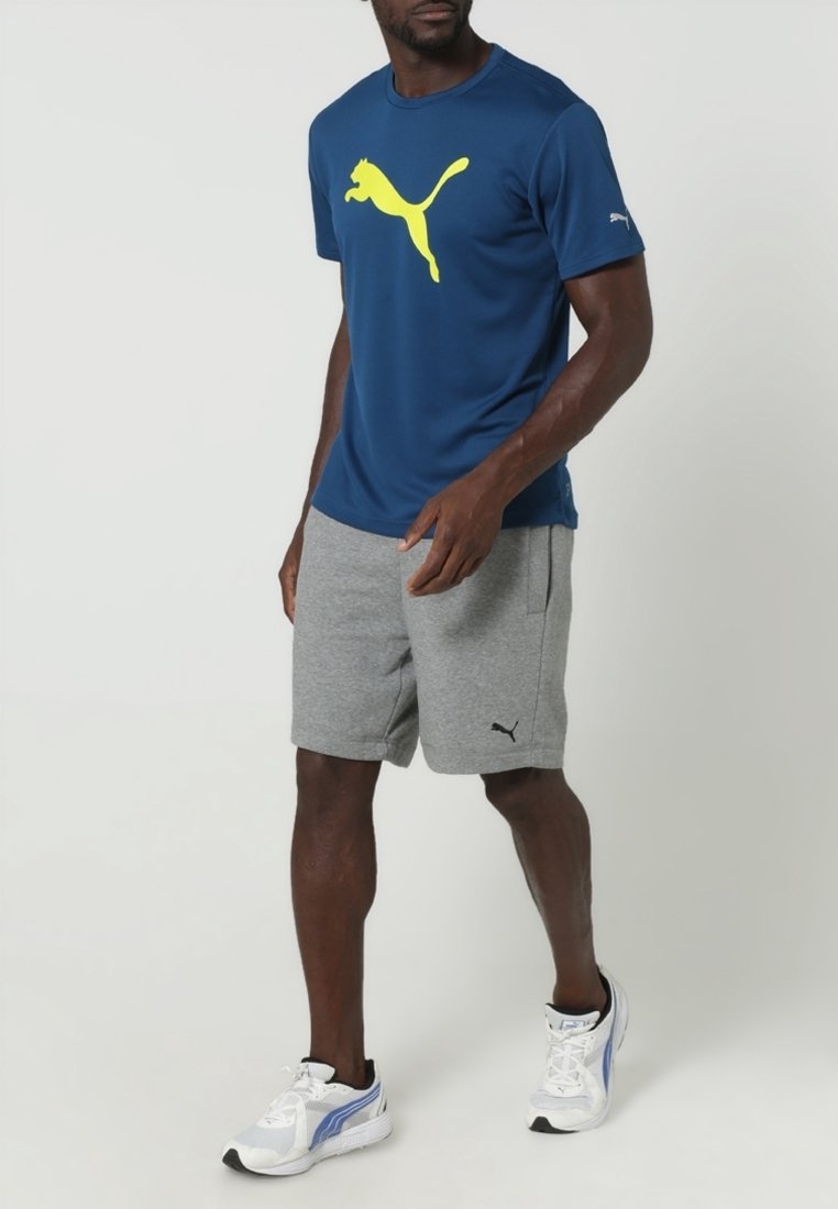 Marinblå sportströja med en stor gul Puma-logotyp, ihop med grå shorts som har en svart Puma-symbol, och vita träningsskor.
