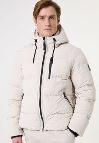 Beige pufferjack met een capuchon, voorzien van zwarte ritsen en accenten. Het materiaal lijkt soepel met gestikte secties voor isolatie.