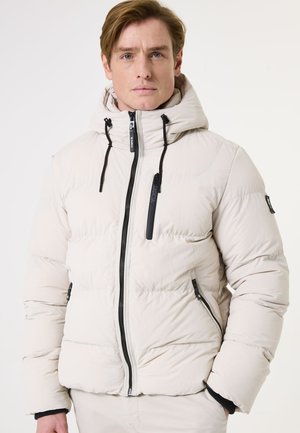 Beige pufferjack met een capuchon, voorzien van zwarte ritsen en accenten. Het materiaal lijkt soepel met gestikte secties voor isolatie.