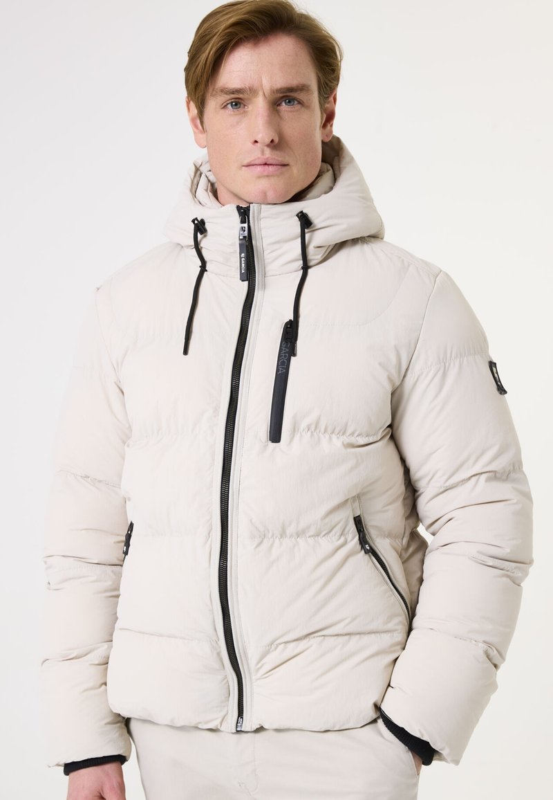 Beige pufferjack met een capuchon, voorzien van zwarte ritsen en accenten. Het materiaal lijkt soepel met gestikte secties voor isolatie.