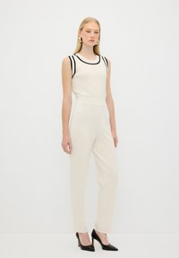 Top sans manches couleur crème avec des accents noirs, associé à un pantalon crème ajusté et des talons hauts noirs, présentant un design épuré et minimaliste.