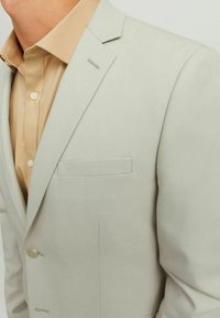 Vaalea harmaa bleiseri, jossa on slim fit -leikkaus, yksittäinen nappisuljenta, uurrettu kaulus ja rintapocket. Sen alla on vaaleanbeige nappikauluspaita.