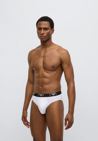 Slip breve bianco con fascia elastica nera brandizzata, design aderente, texture liscia e taglio a vita media. Modello mostra la vista frontale.