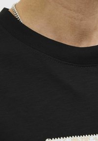 Sort bomulds t-shirt med ribbet halsudskæring og et struktureret nederste hjørne med et kontrasterende beige zigzag-mønster. Sølvkæde synlig.