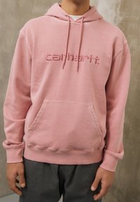 Růžová mikina s kapucí s klokaní kapsou, stahovací šňůrkou a vyšitým logem "carhartt". Materiál působí měkce a má hladkou texturu.