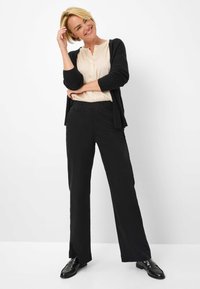 Zwarte wijde broek met een gladde textuur, gecombineerd met een lichtbeige shirt en een donkere cardigan. Zwarte instappers maken de outfit compleet.