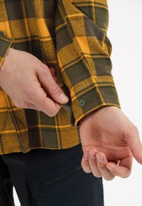 Flanellen shirt met een gele en olijfgroene ruitpatroon, met knoopmanchetten en een zachte textuur, weergegeven met een hand die de mouw aanpast.